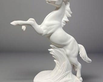 May include: White ceramic sculpture of a horse rearing up on its hind legs, with its front legs raised in the air. The horse is standing on a base with a textured surface that resembles grass.