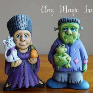 May include: Two hand-painted ceramic figurines, one depicting a woman with a cat and the other a green monster holding a frog. The woman is wearing a purple dress and a gray hat. The monster is wearing a blue shirt and gray pants. The text "Clay Magic Inc." is visible in the background.