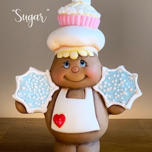 May include: A gingerbread person with a white apron and a red heart on the front. The gingerbread person is wearing a white cupcake hat with pink frosting and a pink candy on top. The gingerbread person has blue eyes and a brown face. The gingerbread person is holding two blue and white snowflake wings. The text "Clay Magic Inc." and "Sugar" is visible in the background.