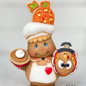 May include: A ceramic figurine of a gingerbread person wearing a pumpkin hat and an apron, holding a pie and a turkey. The figurine is brown, white, and red with a heart on the apron. The turkey is wearing a pilgrim hat. The figurine is labeled "4238".