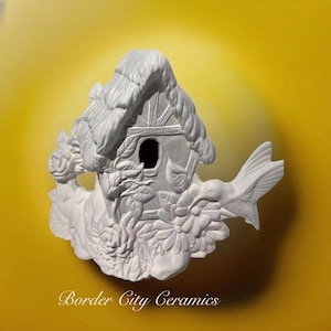 May include: White ceramic birdhouse with a thatched roof and a hummingbird in flight. The birdhouse is decorated with flowers and leaves. The text "Border City Ceramics" is visible at the bottom of the image.