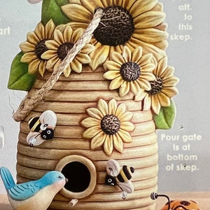 May include: A ceramic beehive with a bluebird perched on the edge. The beehive is decorated with sunflowers and bees. The beehive is 10.5 inches tall and 7.25 inches wide. The text on the image reads: "4313 $84 Lg. Bee Skep 10.5"T x 7.25"W"