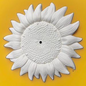 May include: A white, circular, sunflower-shaped wall plaque or decorative object with a textured, seed-like center and radiating petals. The piece features a small, dark, circular hole in the exact center of the flower head, providing a focal point.