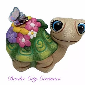 May include: A ceramic turtle figurine with a green shell and a flower design. The turtle has large blue eyes and a pink nose. A butterfly with iridescent wings is perched on the turtle's back.