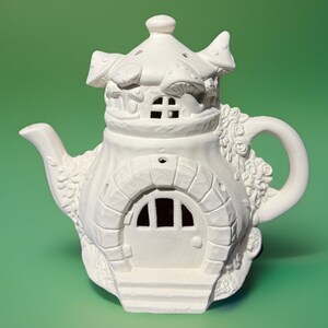 May include: A white ceramic teapot shaped like a fairy house. The teapot features a mushroom-shaped roof, a stone arch doorway, and a handle adorned with floral details. The design includes a small window and a spout.