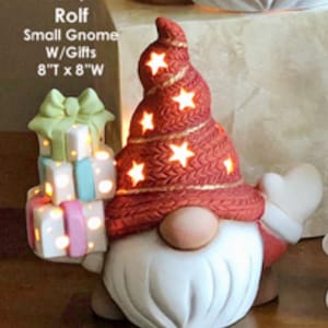 May include: A ceramic gnome figurine with a red knit hat adorned with star-shaped cutouts. The gnome holds a stack of lit gift boxes. The text on the image reads "Rolf Small Gnome W/Gifts 8"T x 8"W".