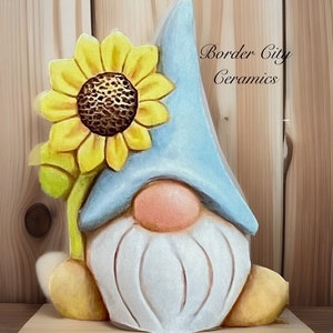 May include: A ceramic gnome figurine with a blue hat and a yellow sunflower on top. The gnome has a white beard and a pink nose. The figurine is painted in a whimsical style. The text "Border City Ceramics" is visible in the background.