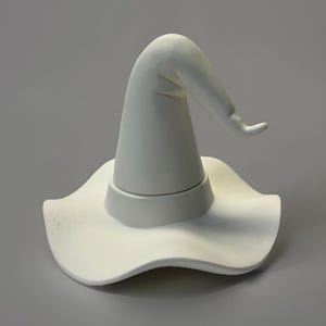 May include: A white ceramic witch hat with a wide, wavy brim and a curved, pointed top. The hat is a solid, matte white color and appears to be handmade. The design is simple and elegant, suitable for Halloween or other themed decor.