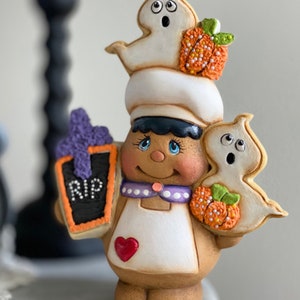 May include: A gingerbread person dressed as a baker holding a tombstone cookie that says "RIP" and two ghost cookies, one with a pumpkin. The gingerbread person is wearing a white apron with a red heart and a purple and white polka dot collar.