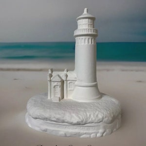 May include: White ceramic lighthouse figurine with a small house attached to the base. The lighthouse has a detailed design and is sitting on a base that looks like a rocky shore. The base has the text "Est. 1898" on it.