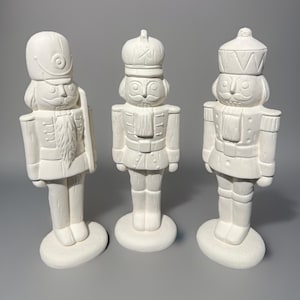 May include: Three unpainted ceramic nutcracker figurines. Each figure is in a different pose, with details like a mustache, uniform, and hat. The figurines are on oval bases and are ready to be painted.