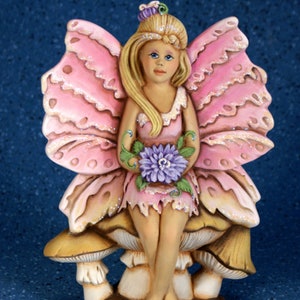 May include: A pink fairy figurine with butterfly wings sits on a mushroom. The fairy is holding a purple flower and has blonde hair and a pink dress.