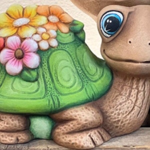 May include: A ceramic turtle figurine with a green shell adorned with colorful flowers. The turtle has a smiling face, large blue eyes, and detailed textured skin. The figurine is a decorative garden or home accent.