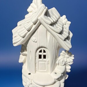 May include: A white ceramic fairy house with a textured roof, door, and decorative floral accents. The house has a chimney and a small door with a cross-shaped window. The overall design is whimsical and detailed.
