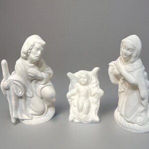May include: White ceramic figurines of the nativity scene. The figures are of Mary, Joseph, and baby Jesus. The figures are all standing on a white base.