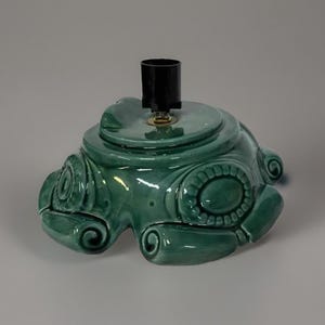 May include: A green ceramic Christmas tree stand with a decorative design. The stand has a metal base and a black plastic socket for a light.