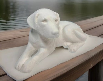 Könnte beinhalten: Eine weiße Keramikskulptur eines liegenden Hundes. Die Skulptur steht auf einer Holzoberfläche mit einem verschwommenen Hintergrund eines Sees.