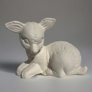 May include: A ceramic figurine of a fawn in a resting pose. The off-white sculpture features detailed textures and rounded forms, including the fawn's ears, body, and paws. The figurine is designed to be painted.