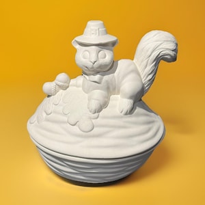May include: A white ceramic squirrel figurine wearing a pilgrim hat and bow tie, perched atop a large walnut-shaped container. The squirrel holds an acorn. The container has a textured, ridged design. The background is a solid yellow.