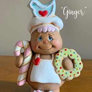 May include: Gingerbread person figurine with a white chef hat, holding a candy cane and a wreath. The figurine is decorated with colorful details and has a blue bird on top of the hat. The text "Ginger" is written on the image.