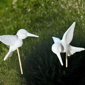 May include: Two white hummingbird garden stakes. Each bird is sculpted with detailed wings and a long beak, mounted on a wooden dowel. The stakes are set against a backdrop of green grass and foliage, suggesting outdoor use for garden decor.