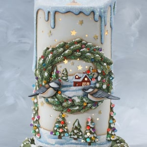 May include: A ceramic candle holder shaped like a candle with a winter scene. The candle is decorated with a wreath, two birds, and a small house. The base of the candle holder is shaped like a snow-covered hill.