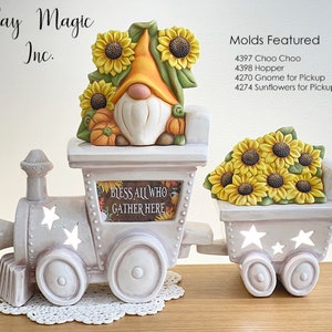 May include: A white ceramic train with a gnome and sunflowers on the front car. The train has a sign that reads "Bless All Who Gather Here". The train is a light gray color with white stars on the sides. The train is on a wooden surface.