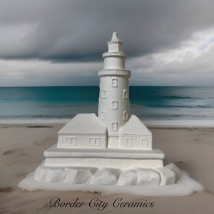 May include: White ceramic lighthouse figurine with a detailed design, sitting on a sandy beach with a blue ocean in the background. The figurine has a tower with windows and a small house on each side. The text "Border City Ceramics" is visible at the bottom of the image.