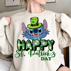 Pode incluir: Camisola bege com um personagem de desenho animado azul usando um chapéu verde com um trevo. As palavras "HAPPY St. Patrick's DAY" são impressas em verde com detalhes de trevo. Roupas temáticas do Dia de São Patrício.