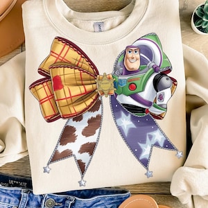 May include: A beige sweatshirt featuring a cartoon image of Buzz Lightyear from Toy Story. The image shows Buzz in his space ranger uniform with a large bow made from plaid fabric, cow print fabric and purple fabric with white stars.