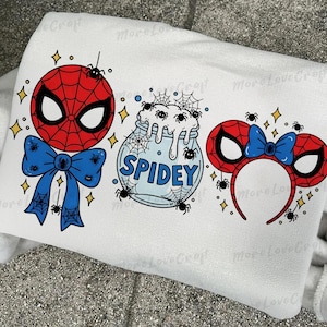 May include: White sweatshirt with three Spider-Man designs. These include a Spider-Man mask with a blue bow, a jar with the word "SPIDEY", and Spider-Man ears with a blue bow. The designs are red, blue, and black.