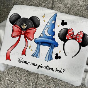 Op de afbeelding: Witte sweatshirt met illustraties van een zwarte Mickey Mouse-hoed met een rode strik, een blauwe tovenaars hoed met sterren en een Minnie Mouse-haarband met een rode polka dot strik. De tekst "Some imagination, huh?" staat eronder.