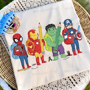 May include: A cream-coloured t-shirt featuring a graphic print of four superheroes dressed for school. Spider-Man, Iron Man, Hulk, and Captain America are illustrated carrying backpacks and holding school supplies like pencils and books.