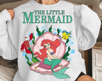 Puede incluir: Sudadera blanca con un estampado gráfico de Ariel de La Sirenita sentada en una concha. El texto "THE LITTLE MERMAID" está impreso en letras verdes en la camiseta.
