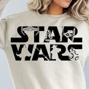 May include: A beige sweatshirt with a black "STAR WARS" graphic. The graphic includes silhouettes of characters and spaceships from the Star Wars franchise.