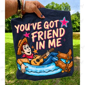 May include: Navy blue t-shirt featuring the text "YOU'VE GOT FRIEND IN ME" with a cartoon of Woody from Toy Story playing a guitar. Woody is wearing a cowboy hat, red bandana, blue jeans, and brown boots. The design includes pink and blue stars.