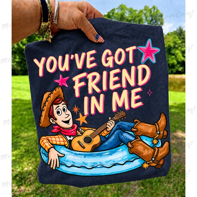 May include: Navy blue t-shirt featuring the text "YOU'VE GOT FRIEND IN ME" with a cartoon of Woody from Toy Story playing a guitar. Woody is wearing a cowboy hat, red bandana, blue jeans, and brown boots. The design includes pink and blue stars.