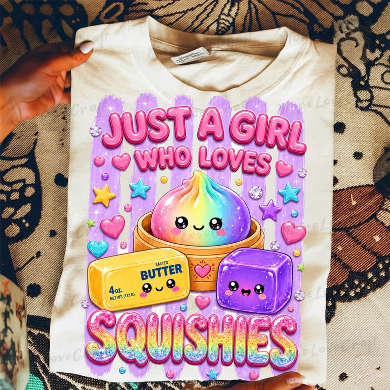 Puede incluir: Una camiseta color crema con un gr&aacute;fico que incluye el texto JUST A GIRL WHO LOVES SQUISHIES. El dise&ntilde;o muestra una bola de masa arco&iacute;ris en una vaporera de bamb&uacute;, una barra de mantequilla salada amarilla marcada con 113g y un cubo morado.