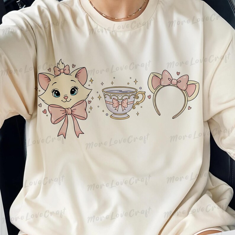 May include: Cream-colored long-sleeve shirt featuring a cartoon illustration of a cat, teacup, and headband with bows. The cat has a pink bow and the teacup has a pink bow. The headband has cat ears and a pink bow. The design includes hearts and stars.