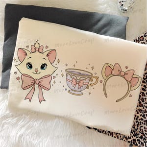 May include: Cream-colored pillowcase featuring a cartoon cat face with a pink bow, a teacup with a pink bow, and a cat ear headband with a pink bow. The design includes hearts and stars. The pillowcase is on a white surface.