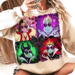 May include: Beige sweatshirt featuring a colorful graphic of four cartoon villains wearing heart-shaped sunglasses and blowing bubblegum bubbles. The graphic includes Cruella de Vil, Ursula, Maleficent, and the Evil Queen. A black bow is in the center of the graphic.