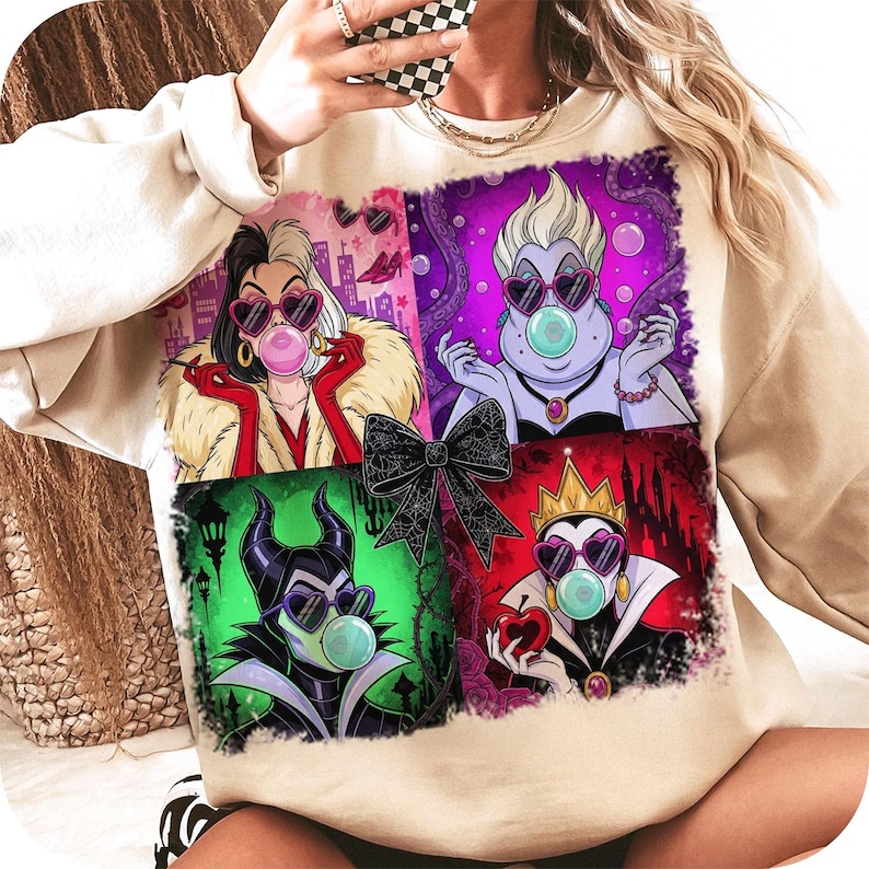 May include: Beige sweatshirt featuring a colorful graphic of four cartoon villains wearing heart-shaped sunglasses and blowing bubblegum bubbles. The graphic includes Cruella de Vil, Ursula, Maleficent, and the Evil Queen. A black bow is in the center of the graphic.