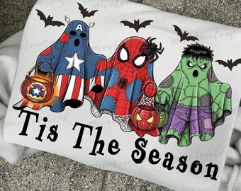 May include: A white sweatshirt with a Halloween design. It features three cartoon ghosts in superhero costumes: Captain America, Spider-Man, and the Hulk, each carrying a pumpkin lantern. The text "Tis The Season" is printed below, with bat silhouettes above.
