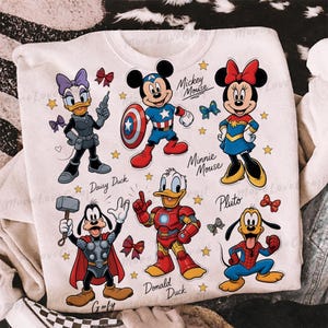 May include: White sweatshirt featuring cartoon characters Mickey Mouse, Minnie Mouse, Donald Duck, Daisy Duck, Goofy, and Pluto dressed as superheroes. The design includes stars and bows, with character names below each figure.
