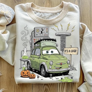 Puede incluir: Sudadera crema con un coche verde de dibujos animados con rasgos de Frankenstein. El dise&ntilde;o incluye una escena de laboratorio, calabazas, rayos y el texto "IT'S A-LUIGI!"