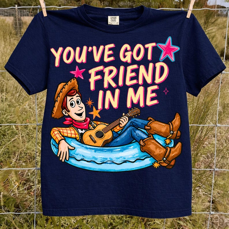 May include: Navy blue t-shirt featuring a cartoon cowboy character playing a guitar while lounging in a blue inner tube. The shirt has the text "YOU'VE GOT FRIEND IN ME" in pink and white. The shirt is hanging on a clothesline.