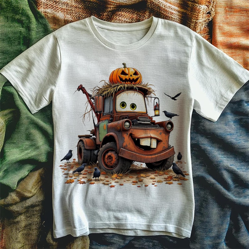 Puede incluir: Camiseta blanca con un dise&ntilde;o de cami&oacute;n de remolque de dibujos animados, coronada con una calabaza tallada. El cami&oacute;n es marr&oacute;n con una cara sonriente y dientes grandes. Cuervos negros y hojas de oto&ntilde;o rodean el cami&oacute;n.