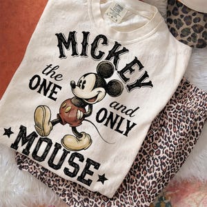 May include: Cream-colored sweatshirt featuring a graphic of Mickey Mouse with the text "Mickey the One and Only Mouse." The design includes a cartoon image of Mickey in his classic outfit. Paired with leopard print pants.