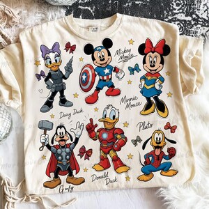 May include: Cream-colored t-shirt featuring cartoon characters Mickey Mouse, Minnie Mouse, Donald Duck, Daisy Duck, Goofy, and Pluto dressed as superheroes. The shirt has the names of the characters printed below them, along with stars and bows.