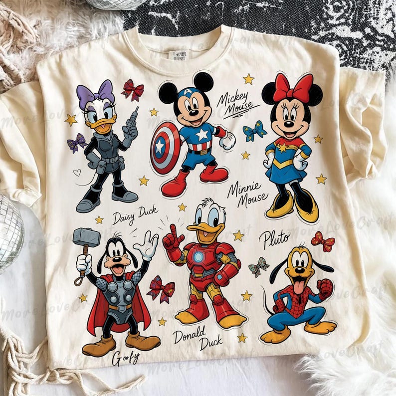 May include: Cream-colored t-shirt featuring cartoon characters Mickey Mouse, Minnie Mouse, Donald Duck, Daisy Duck, Goofy, and Pluto dressed as superheroes. The shirt has the names of the characters printed below them, along with stars and bows.